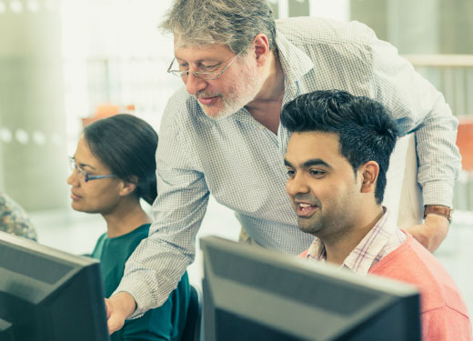 Lesson situation: a young man and a young woman sitting at computers. A teacher is standing behind them explaining something.