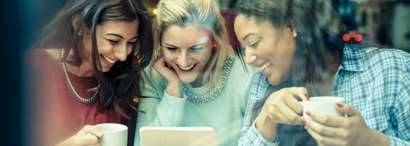 Three women in a café. They are looking at a tablet and laughing together.