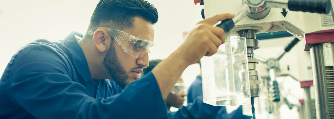 A man wearing safety glasses concentrates while operating a bench drill.