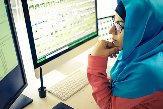 Arbeitssituation: Eine Frau mit Kopftuch sitzt vor zwei Computerbildschirmen.